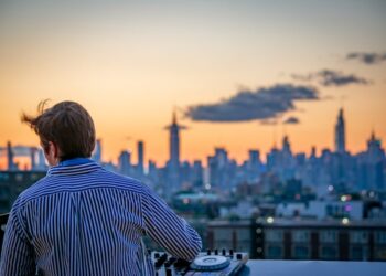 DJ op een rooftopterras die muziek draait terwijl de skyline van de stad achter hem bij zonsondergang gloeit, gekleed in een gestreept shirt.