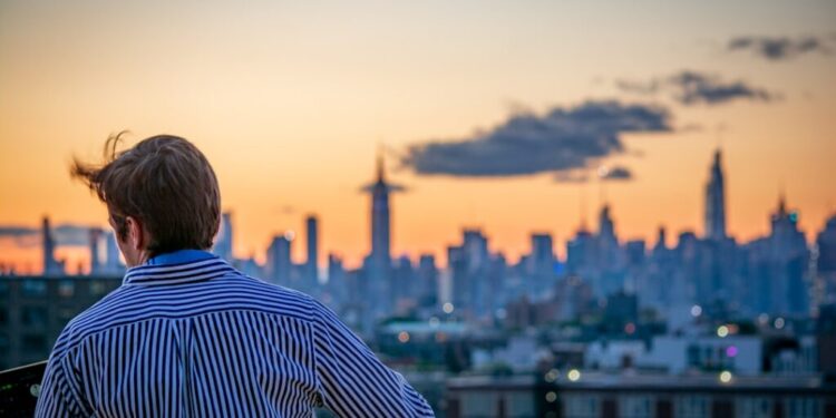 DJ on a rooftop deck mixing music as the city skyline glows at sunset behind him, wearing a striped shirt.