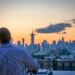 DJ on a rooftop deck mixing music as the city skyline glows at sunset behind him, wearing a striped shirt.