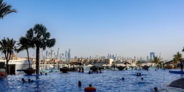 Beach clubs in Dubai worth joining - GUSTOwebzine People swimming in a large outdoor pool with palm trees and a distant city skyline under a clear blue sky.