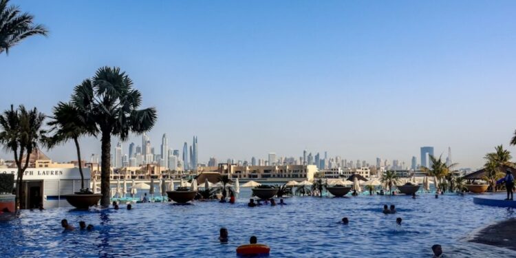 People swimming in a large outdoor pool with palm trees and a distant city skyline under a clear blue sky.