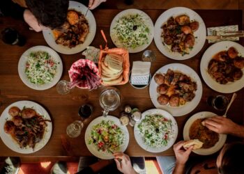 Een groep gasten reikt over een gemeenschappelijke tafel die vol staat met gedeelde schalen, ter ere van de traditie van de gemeenschappelijke tafel.
