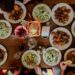 people-sharing-dishes-large-table-unsplash - GUSTOwebzine A group of guests reaching across a communal table covered in shared dishes, celebrating the communal table tradition.