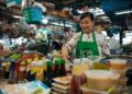 Vendor wearing a green GrabFood apron prepares sauces at a busy market stall with colorful bottles in the foreground.