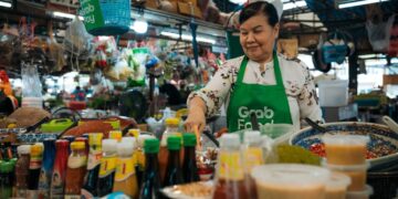 Vendor wearing a green GrabFood apron prepares sauces at a busy market stall with colorful bottles in the foreground.