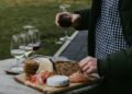 Person wearing a green jacket and checkered shirt holds a glass of red wine over a wooden board with cheese, crackers, bread, and sliced meat outdoors.