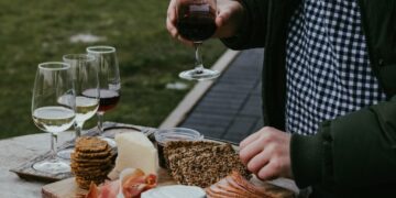 Person wearing a green jacket and checkered shirt holds a glass of red wine over a wooden board with cheese, crackers, bread, and sliced meat outdoors.