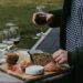 Person wearing a green jacket and checkered shirt holds a glass of red wine over a wooden board with cheese, crackers, bread, and sliced meat outdoors.