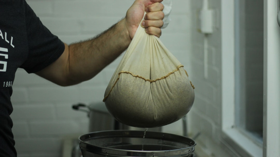 A person straining a batch of drinking vinegars through cheesecloth into a clear glass jar on a wooden counter