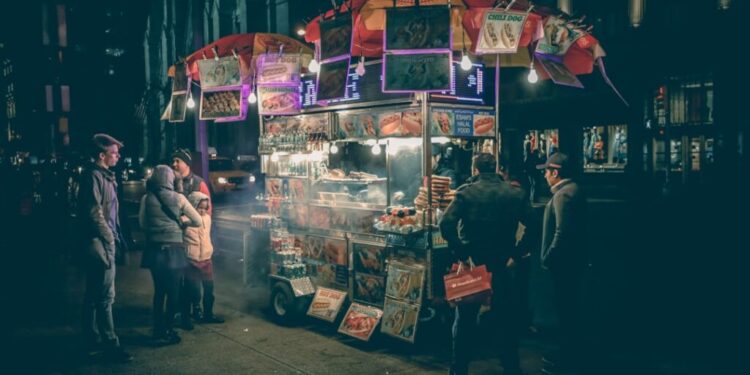 Nighttime street food cart glowing with lights as customers gather around the menu.