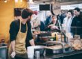 Street food markets London guide - GUSTOwebzine Bearded vendor in a beige apron works at an outdoor market stall, cups stacked beside him, and customers in line.