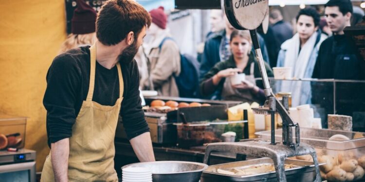Bearded vendor in a beige apron works at an outdoor market stall, cups stacked beside him, and customers in line.
