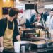 Street food markets London guide - GUSTOwebzine Bearded vendor in a beige apron works at an outdoor market stall, cups stacked beside him, and customers in line.