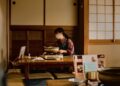 Gluten-free izakaya Tokyo guide - GUSTOwebzine Woman wearing a mask cooks at a low wooden table on a portable stove in a traditional Japanese room with tatami mats and shoji screens.