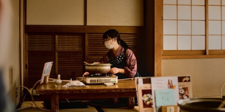 Woman wearing a mask cooks at a low wooden table on a portable stove in a traditional Japanese room with tatami mats and shoji screens.