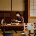 Woman wearing a mask cooks at a low wooden table on a portable stove in a traditional Japanese room with tatami mats and shoji screens.
