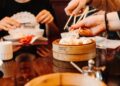 Dim sum London best restaurants guide - GUSTOwebzine People using chopsticks to pick dumplings from a wooden steam basket at a dim sum meal.