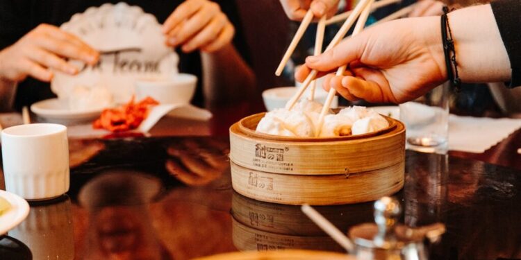 People using chopsticks to pick dumplings from a wooden steam basket at a dim sum meal.
