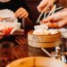 People using chopsticks to pick dumplings from a wooden steam basket at a dim sum meal.