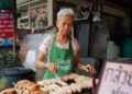 Elderly woman wearing a green GrabFood apron grills skewers at a street food stall.