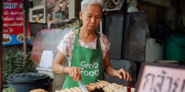 Elderly woman wearing a green GrabFood apron grills skewers at a street food stall.