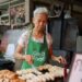 Elderly woman wearing a green GrabFood apron grills skewers at a street food stall.
