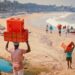 People carrying red plastic crates on a sandy beach with boats along the shore under a sunny sky.