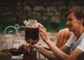 woman-pouring-cold-brew-tea-jar-unsplash - GUSTOwebzine A woman carefully pouring cold brew tea from a glass jar into a cup on a wooden kitchen counter