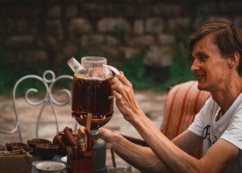 woman-pouring-cold-brew-tea-jar-unsplash - GUSTOwebzine A woman carefully pouring cold brew tea from a glass jar into a cup on a wooden kitchen counter