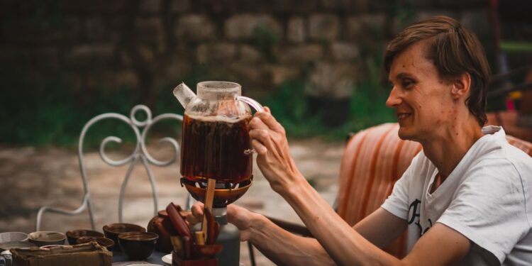 woman-pouring-cold-brew-tea-jar-unsplash - GUSTOwebzine A woman carefully pouring cold brew tea from a glass jar into a cup on a wooden kitchen counter