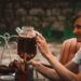 woman-pouring-cold-brew-tea-jar-unsplash - GUSTOwebzine A woman carefully pouring cold brew tea from a glass jar into a cup on a wooden kitchen counter