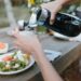 woman-pouring-fruit-shrub-jar-unsplash - GUSTOwebzine A woman carefully pouring homemade drinking vinegars from a glass jar into a small bottle in a sunlit kitchen