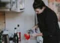 A woman carefully pouring a glass of fermented drinks at a bright kitchen counter, surrounded by seasonal autumn produce