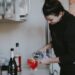 Una mujer vertiendo cuidadosamente un vaso de bebidas fermentadas en una encimera de cocina luminosa, rodeada de productos de temporada de otoño