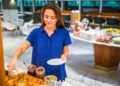 A woman in a blue shirt serves breakfast at a buffet, placing croissants and jars of jam on the table.