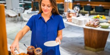 A woman in a blue shirt serves breakfast at a buffet, placing croissants and jars of jam on the table.
