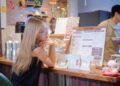 Woman with long blonde hair and glasses leaning on a cafe counter, studying a large menu board.