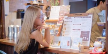 Woman with long blonde hair and glasses leaning on a cafe counter, studying a large menu board.