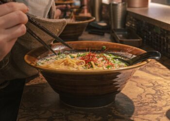 Een kom ramen op een marmeren tafel, gegarneerd met groenten en rode chilidraadjes, waarbij stokjes noedels omhoog tillen en een lepel in de kom rust.
