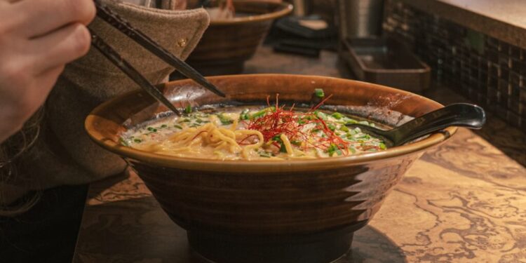 Een kom ramen op een marmeren tafel, gegarneerd met groenten en rode chilidraadjes, waarbij stokjes noedels omhoog tillen en een lepel in de kom rust.
