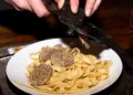 Fettuccine pasta on a white plate with thin truffle shavings being shaved over it by a hand-held slicer.