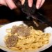Fettuccine pasta on a white plate with thin truffle shavings being shaved over it by a hand-held slicer.