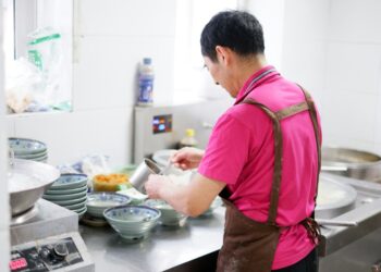 Ein Koch baut vorsichtig eine aromatische Basis in einem Topf auf und bereitet ein klassisches Reis-Eintopfgericht mit geschichteten Gewürzen und Fond zu.