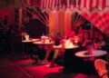Dineren op een terras in Dubai 6 plekken om nu te reserveren - GUSTOwebzine Groep vrienden gezeten aan ronde buitentafels onder een gestreepte luifel, drankjes en bloemen op tafel in roze avondlicht.
