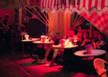 Groep vrienden gezeten aan ronde buitentafels onder een gestreepte luifel, drankjes en bloemen op tafel in roze avondlicht.