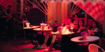 Dineren op een terras in Dubai 6 plekken om nu te reserveren - GUSTOwebzine Groep vrienden gezeten aan ronde buitentafels onder een gestreepte luifel, drankjes en bloemen op tafel in roze avondlicht.