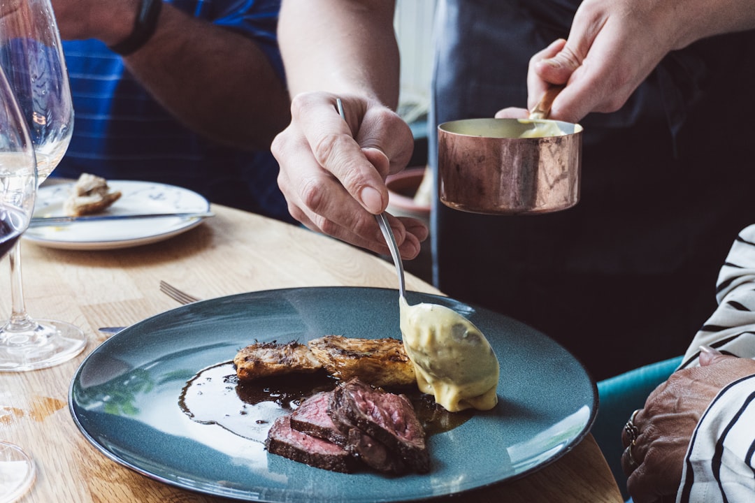 A chef pouring a finished pan sauce over a protein on a white dinner plate in a modern kitchen