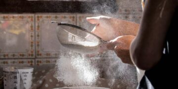 Eine Person siebt in einer Küche Mehl durch ein Metallsieb in eine Schüssel, wobei Mehlstaub in der Luft schwebt.