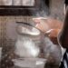 Person sifts flour through a metal sieve into a bowl in a kitchen, flour drifting in the air.