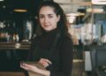 Restaurantmenu's in Singapore in september - GUSTOwebzine Barista met een zwart schort en coltrui staat in een modern café, houdt een notitieboekje vast en glimlacht naar de camera.
